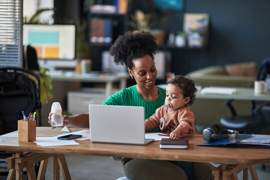 Mother multitasking by using laptop while holding baby in home office. Casual setting with productivity tools and infant care essentials visible