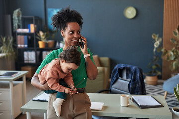 African American woman holding her baby while multitasking on phone managing work in modern office