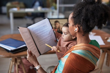 Mother multitasking by preparing documents while watching over child in home office. Child engaging...