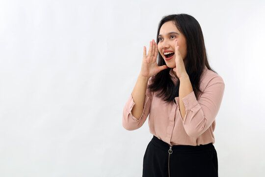 Young Asian woman shouting to announce information to empty space beside, looking enthusiastic and happy facing sideways, wearing shirt, standing isolated on white background.