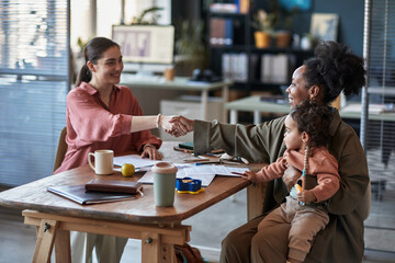 Caucasian woman shaking hands with African American woman holding a child in modern office setting...
