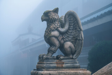 A stone statue of a ferocious griffin. A statue of a mythical animal standing in a misty garden