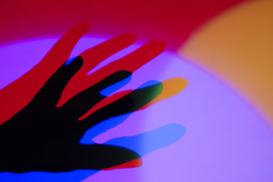 A human hand casts a dramatic shadow against a vibrant background of overlapping red, blue, and green lights. 