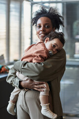 Portrait of woman holding baby, both looking at camera with soft expressions in modern office...