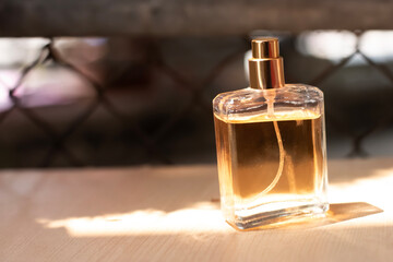 A glass bottle of perfume, square, with brown water, placed on a light brown wooden background, with a blurred background of iron mesh, with sunlight shining through.