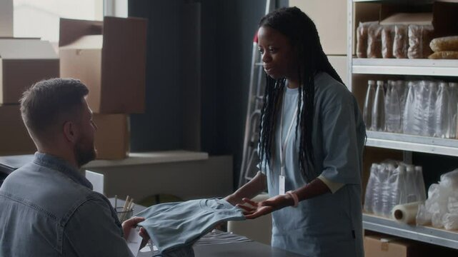 Medium long shot of female African American coordinator giving uniform and ID badge to male newcomer during orientation at donation center stocked with food supplies