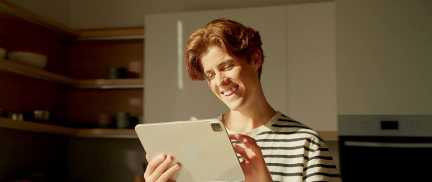 Easygoing young Caucasian man in striped T-shirt holding digital tablet and touching screen. Guy reading funny messages from friend and laughing. Tracking moving around. 360 degree kitchen. Parallax