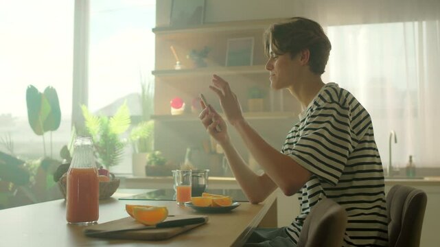 360 degree shot. Footage of male Caucasian student holding smartphone and calling via video call. Happy guy shrugging shoulders and talking at breakfast table with fresh ingredients in kitchen