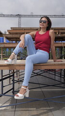 Stylish young Asian woman in a red top and blue jeans sits on a wooden bench on outdoor stands, showcasing a modern, casual look with platform sandals