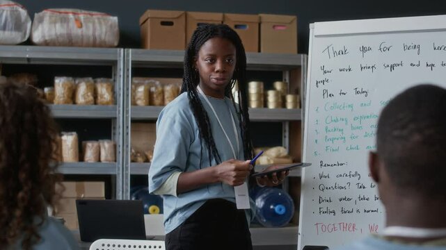 Handheld shot of female African American coordinator discussing work with team in volunteers center with food supplies