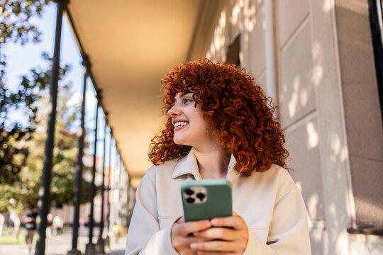 Student with curly red hair smiling while holding a smartphone on a university campus
