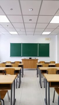 Empty classroom shows rows of wooden desks, green chalkboard, teacher's desk, and white tiled floor for elementary education.