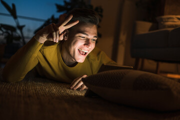 Cheerful man gesturing peace sign on video call while lying on carpet in living room during night