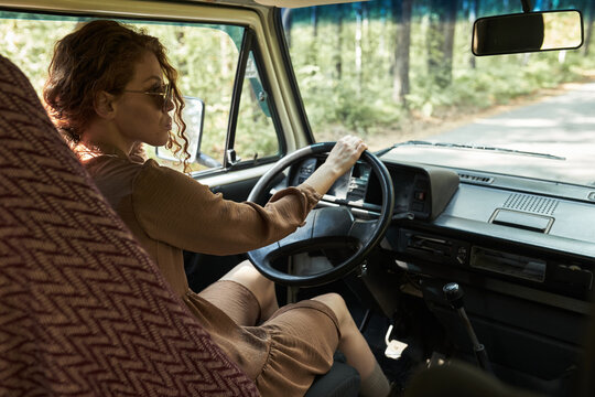 Woman sitting behind wheel of vintage car, driving through forest road with sunlight illuminating interior, wearing casual attire and sunglasses