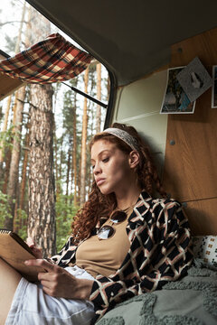 Woman sitting inside a camper van, holding notebook and pen while writing. Scenic forest visible through open door, providing tranquil background