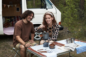 Smiling couple cooking meal together at campsite near van, enjoying outdoor adventure. Picnic table with various utensils and cooking gear on display in wooded area