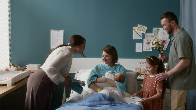 Happy family visting young mother and newborn baby in maternity hospital, giving her flowers, postcard and gift