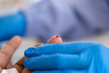 Close-up of a healthcare worker in blue gloves holding a patient's finger with a drop of blood...