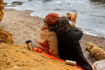 Two women sit closely on the beach, sharing a cozy moment with coffee and pastries, wrapped in...