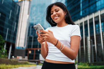 A young woman is smiling broadly while she is using her smartphone in an urban setting
