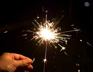 A hand holds a burning sparkler at night, creating bright sparks and light trails against a dark background.