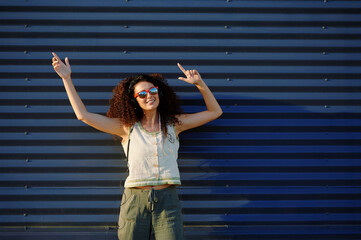 joyful woman with sunglasses raises arms against corrugated metal wall, embracing summer vibes with...