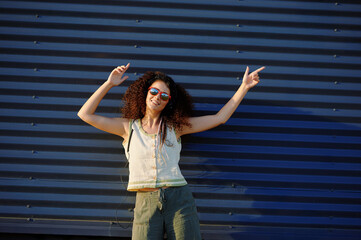 woman wearing sunglasses dancing joyfully against corrugated blue wall. fashionable casual attire...