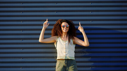woman with curly hair wearing sunglasses gestures enthusiastically against metal wall. casual style...