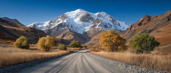 Mountain Road in Autumn: A gravel road winds through a valley towards a majestic snow-capped mountain, flanked by autumnal trees in vibrant yellow and green hues under a clear blue sky. 