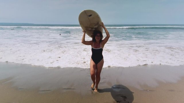 Active mature woman in wetsuit with surfboard at the beach, enjoying a healthy lifestyle. Old senior female doing surfing on Bali. Elderly lady holding surf board. Tropical coast in asia, big wave.