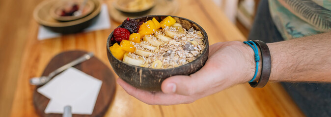 Banner of unrecognizable chef hand showing exotic vegan breakfast made with mango, banana, raspberries, cereals and muesli in coconut bowl. Healthy vegetarian dishes concept.