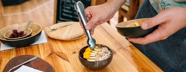 Banner of unrecognizable chef hands preparing a vegan breakfast made with mango, banana, raspberries, cereals and muesli using coconut bowl and metal tongs. Healthy vegetarian dishes concept.