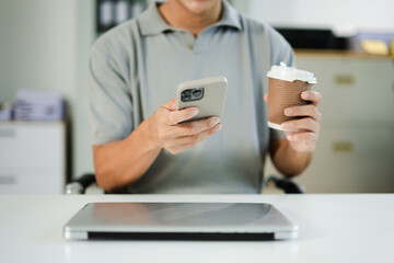 Asian man holding coffee and smartphone, working in modern office with laptop. Perfect for lifestyle