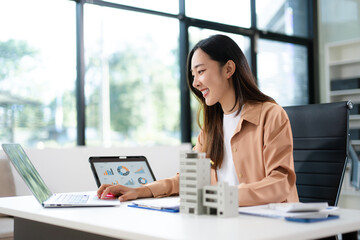 Asian woman analyzing sales data on tablet with real estate model on desk. Ideal for finance, property investment, marketing, and smart business strategy concepts.