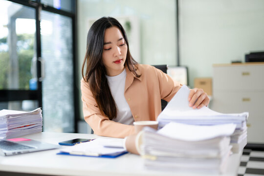 Asian businesswoman focused on sorting stacks of paperwork at her desk with a laptop and coffee cup.