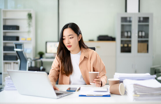 Asian businesswoman focused on sorting stacks of paperwork at her desk with a laptop and coffee cup.