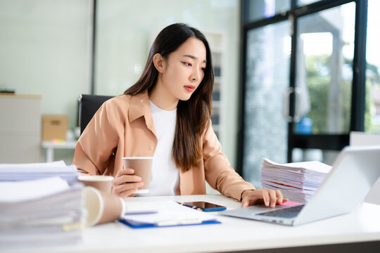 Asian businesswoman focused on sorting stacks of paperwork at her desk with a laptop and coffee cup.