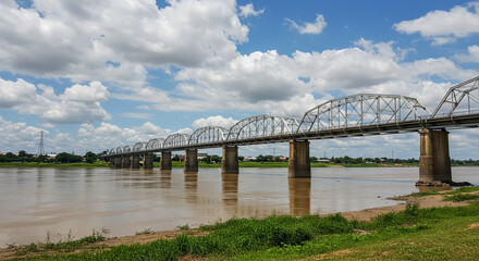 Obraz premium The steel truss bridge spans a wide river under a cloudy blue sky, with trees along the banks.