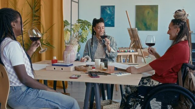 Happy young Caucasian girl in wheelchair clinking wine glasses with African American and biracial girlfriends sitting at table and enjoying paint and sip art class