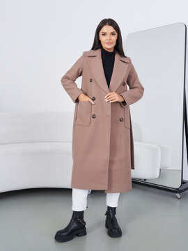 A woman in a long brown coat stands confidently, hands in pockets. She wears a black top, white pants, and chunky black boots. A large mirror and a white sofa are in the room
