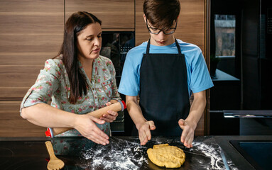 Mother and teenage son preparing dough in kitchen. Woman with rolling pin helping child cooking.