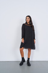 Woman stands in a black polka dot dress with long sleeves and lace collar. She wears black ankle...