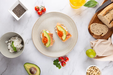 Top view of whole grain toasts with avocado, salmon and cherry tomatoes on white marble with fresh ingredients and glass of kombucha. Concept of healthy food and minimalist aesthetics