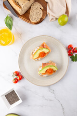 Toasts with avocado, salmon and cherry tomatoes on beige plate. Top view on light marble background with fresh ingredients. Concept of healthy breakfast and balanced lifestyle