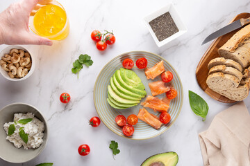 Top view of nutritious breakfast with avocado, salmon, whole grain bread, ricotta, nuts and a glass of kombucha held by a man's hand. Concept of healthy eating and homemade cuisine