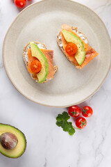 Top view close-up of two toasts with avocado, salmon and cherry tomatoes on beige plate. White marble background. Concept of healthy breakfast and balanced nutrition