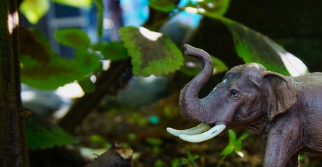 close up of a small plastic elephant toy in the photo with a background of leaves in the park, and it looks like a real elephant with its long trunk