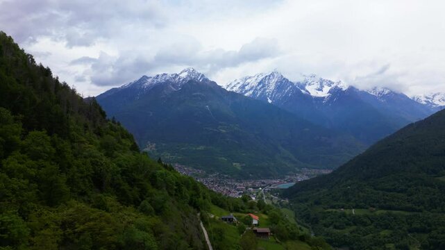 Sweeping aerial view of green forested hills and dramatic snow-covered Alpine peaks under a cloudy sky. Shot near Edolo, Italy Italia