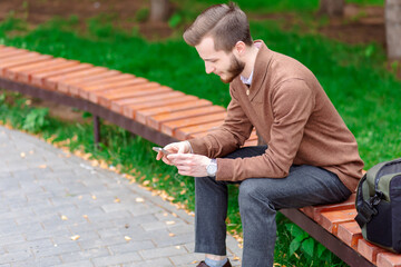 Fototapeta premium Young man with a phone in his hands sits on a bench in a park. Man sitting on bench