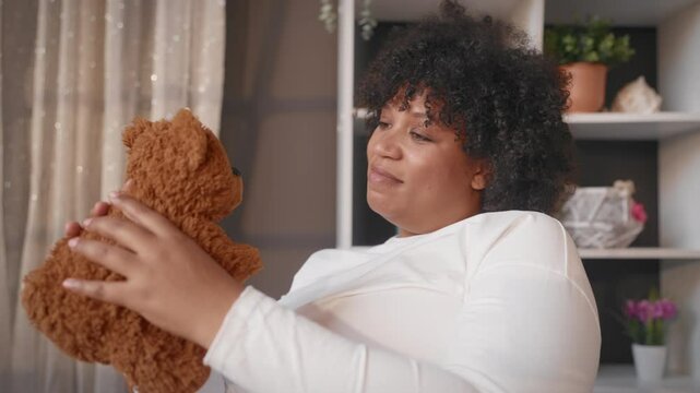 Charming pregnant woman playing with cute teddy bears, toys for future baby. African american black lady resting alone in cozy apartment, healthy pregnancy, good mood and positivity, happy parenthood